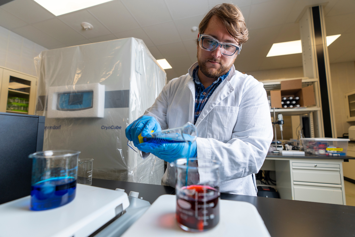 person in lab gear pouring blue liquid from small beaker into a larger beaker that has red liquid