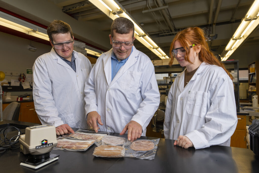 A male student and female student stand on either side of a professor in a lab.