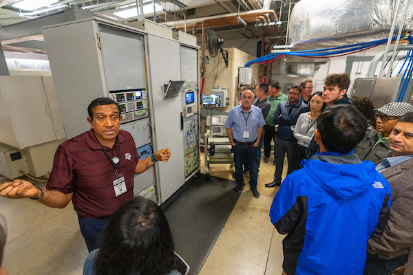 A man speaking to a group of people at the National Center for Electron Beam Research. There is various equipment throughout the room