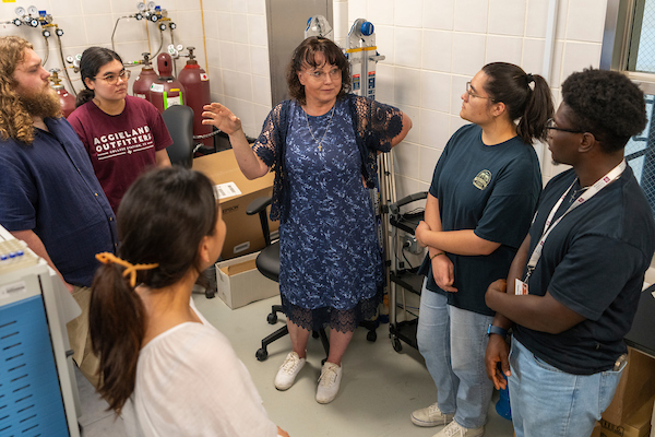 A woman in a blue dress instructs five students in a lab setting. She is standing in the between them and there is various lab equipment behind her.